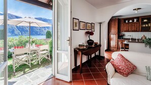 Uma porta aberta dá acesso a uma sala de jantar com mesa e cadeiras, uma cozinha com armários de madeira, e uma vista para montanhas através da porta aberta.