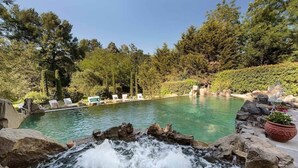 Una piscina con cascada, rodeada de árboles y sillas de descanso.