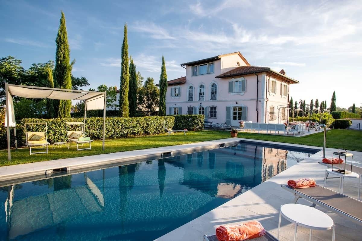Ein Swimmingpool mit Liegestühlen und einer Pergola, umgeben von einem gepflegten Rasen und hohen Bäumen.