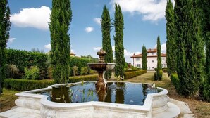 Una fuente en un jardín con árboles altos y un edificio al fondo.