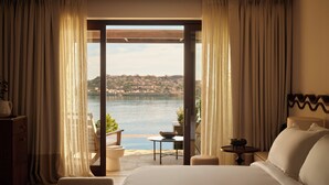 A hotel room with a large bed, a view of a body of water, and a balcony with a table and chairs. - Rosewood Blue Palace (Agios Nikolaos)