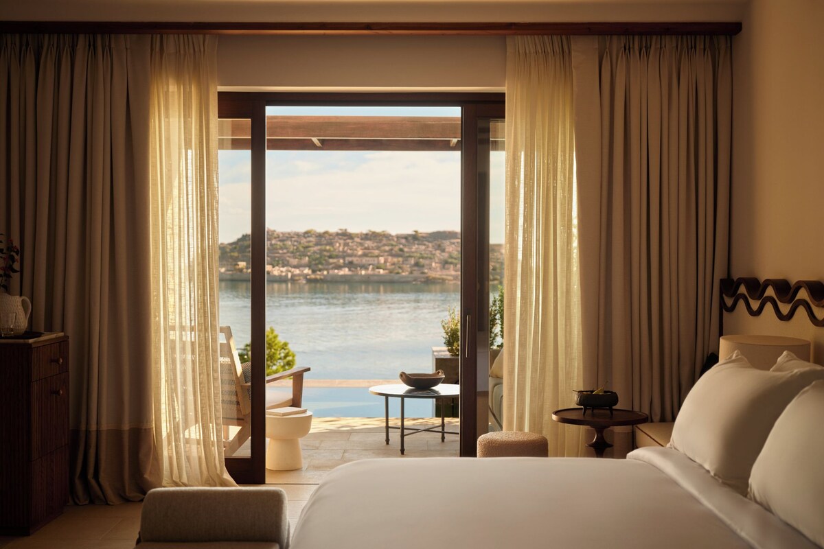 A hotel room with a large bed, a view of a body of water, and a balcony with a table and chairs.