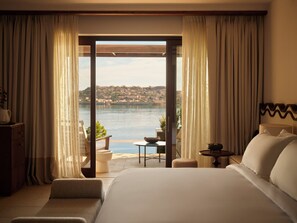 A hotel room with a large bed, a view of a body of water, and a balcony with a table and chairs. - Rosewood Blue Palace (Agios Nikolaos)
