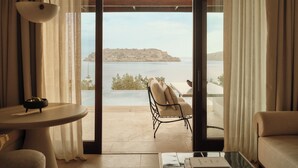A room with a view of a body of water and a distant island, a chair, a table, and a ceiling fan. - Rosewood Blue Palace (Agios Nikolaos)