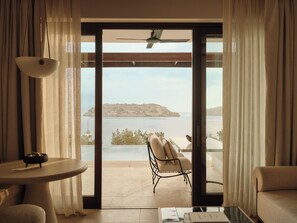 A room with a view of a body of water and a distant island, a chair, a table, and a ceiling fan. - Rosewood Blue Palace (Agios Nikolaos)