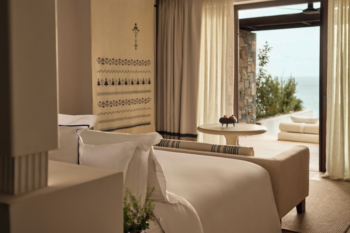 A hotel room with a large bed, a small table, a view of the sea, and a decorative wall.