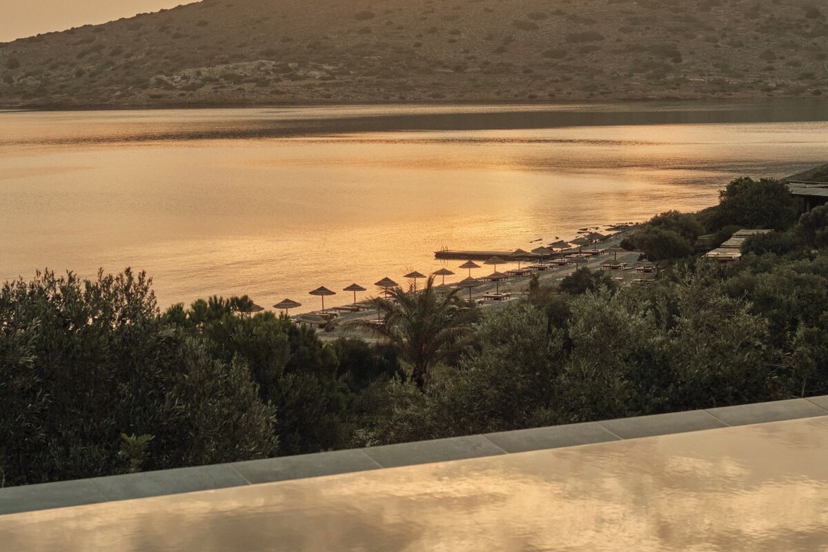 A serene sunset view from a balcony with a pool, overlooking a beach with umbrellas and lounge chairs.
