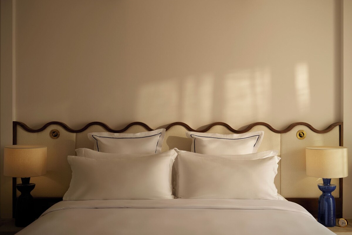 A neatly made bed with white linens and pillows, flanked by two bedside tables with lamps, in a room with a simple, clean design.