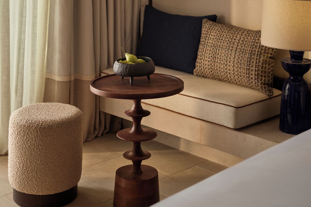 A hotel room with a sofa, a round table with a bowl of fruit, a lamp, and a textured ottoman.