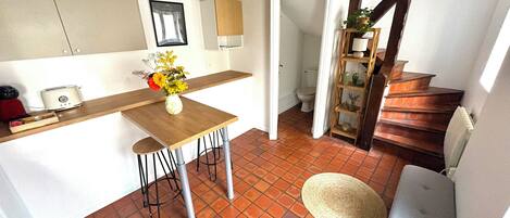 Una cocina con una mesa de madera, taburetes de bar y un jarrón de flores. Se ve una escalera que sube a otra habitación.