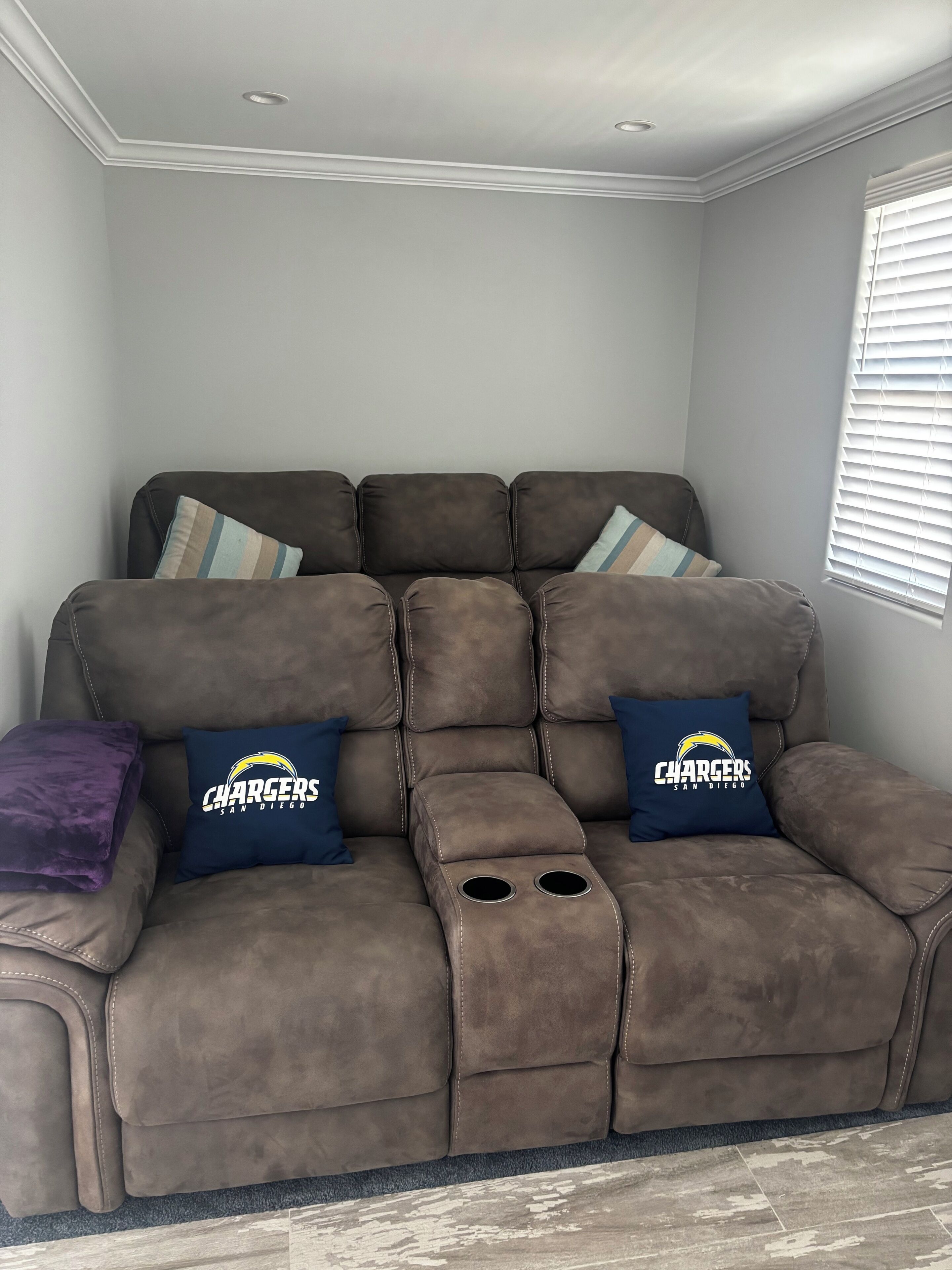 Ein braunes Liegesofa mit zwei blauen Kissen, die das Chargers-Logo zeigen, einem violetten Zierkissen und einem Fenster mit Jalousien.