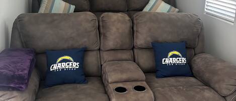Ein braunes Liegesofa mit zwei blauen Kissen, die das Chargers-Logo zeigen, einem violetten Zierkissen und einem Fenster mit Jalousien.