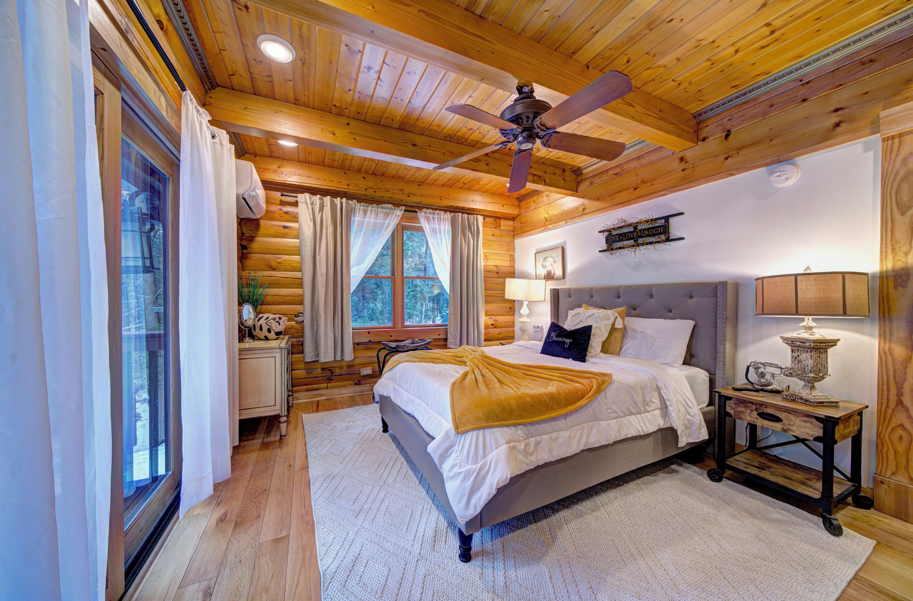 A wooden cabin bedroom with a bed, a nightstand, a ceiling fan, and a window with curtains.