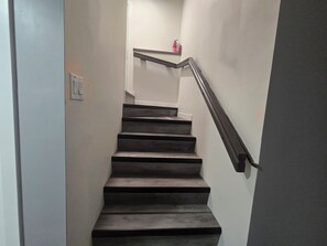 Una escalera con peldaños oscuros y una barandilla metálica, que conduce a un rellano con un extintor.