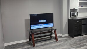 A modern living room with a flat-screen TV on a glass TV stand, grey walls, and a grey carpet. - Sally Room 101-Place in Devonshire Park (Winnipeg)