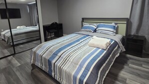 Un dormitorio con cama, espejo, mesita de noche, televisor y luz de techo.