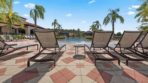 Zona junto a la piscina con sillones de descanso, suelo de baldosas y palmeras.