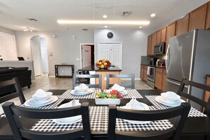 A modern kitchen with a refrigerator, microwave, and oven. There is a dining area with a table set for four, chairs, and a fruit bowl. - Sweet Retreat At Bella Vida (Kissimmee)