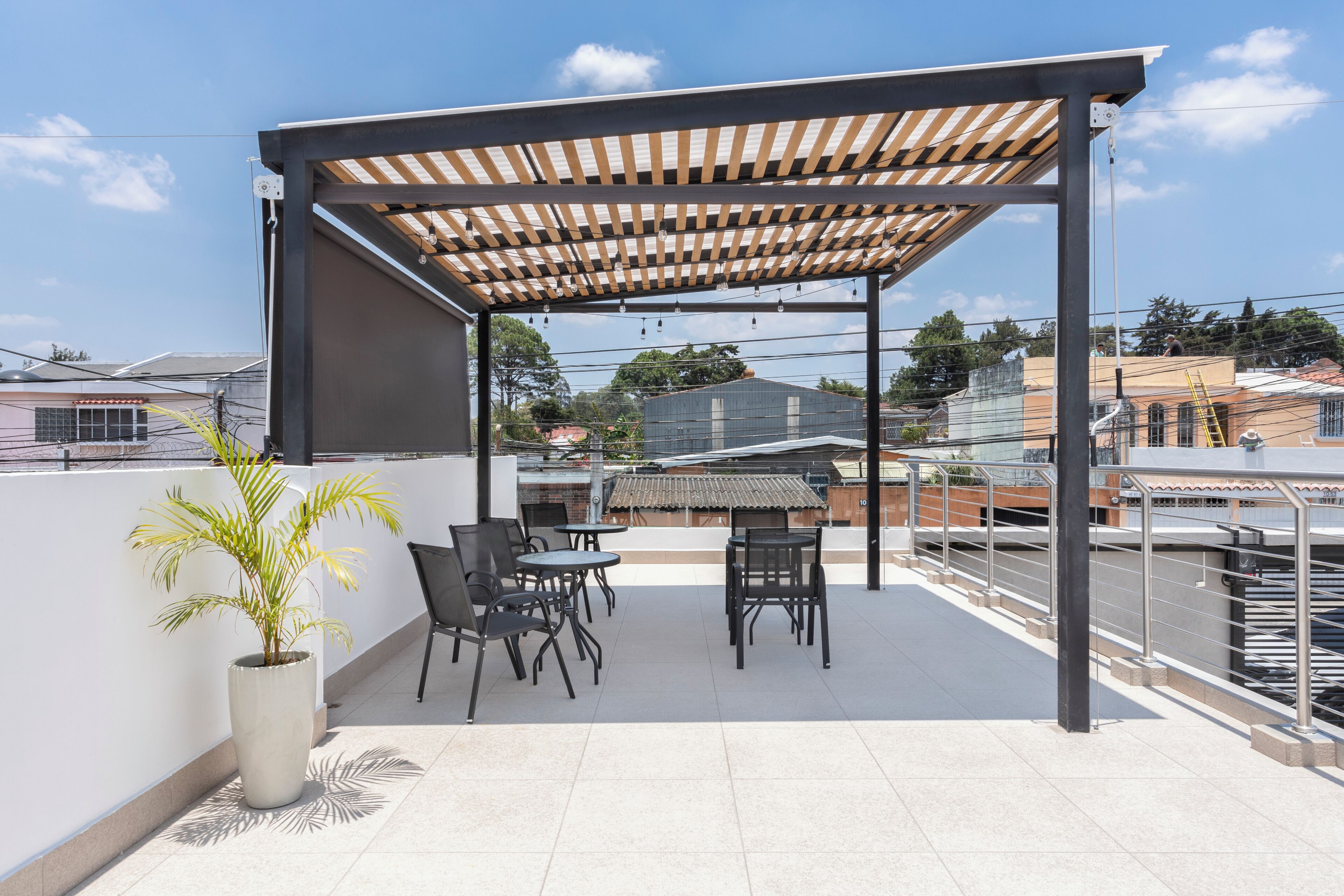 A covered outdoor seating area with tables and chairs, a potted plant, and a view of surrounding buildings.