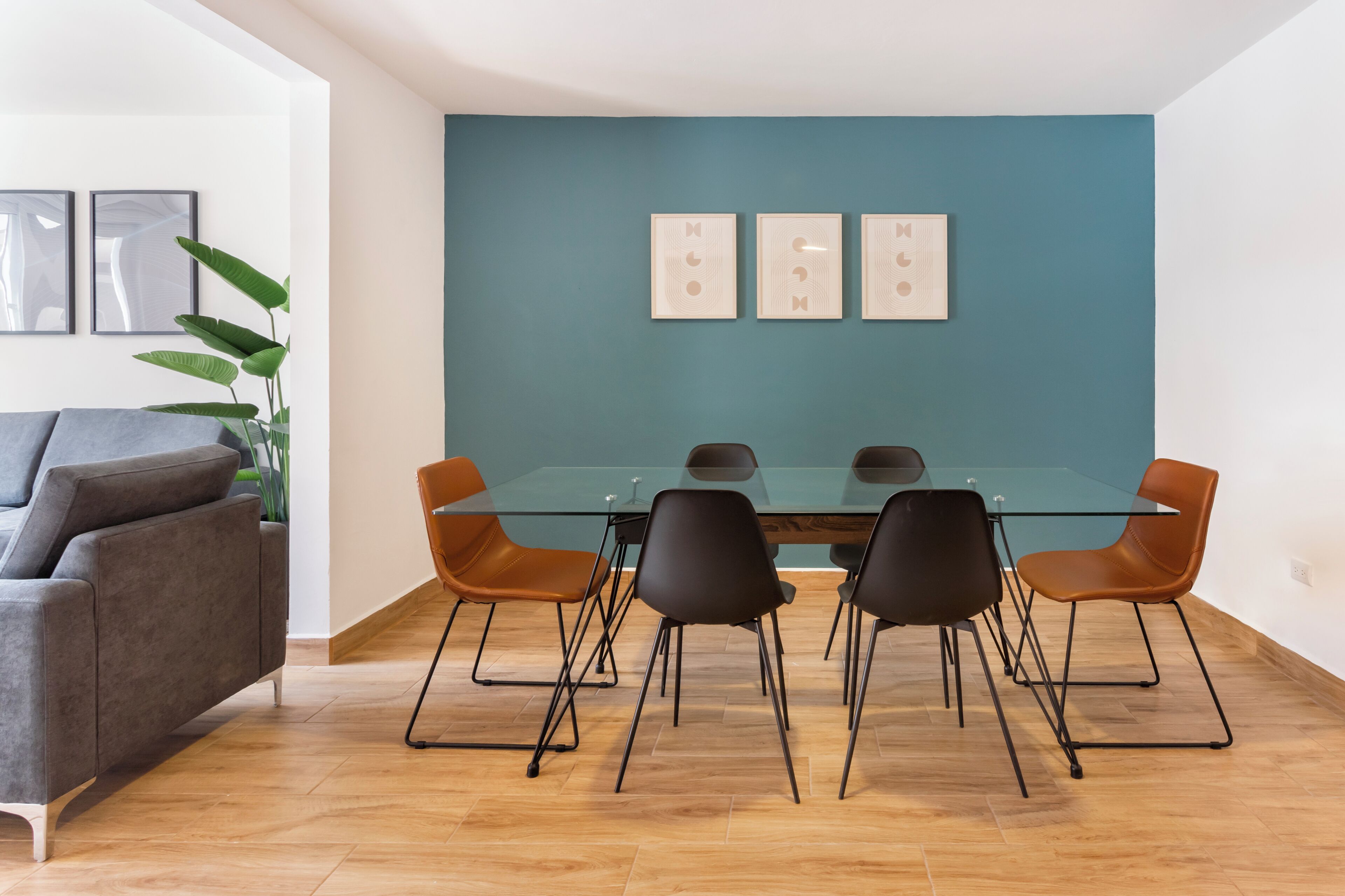 Uma sala de jantar moderna com uma mesa de vidro e cadeiras pretas e castanhas, um sofá cinzento e uma parede de destaque azul-turquesa.