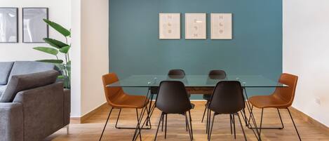 Uma sala de jantar moderna com uma mesa de vidro e cadeiras pretas e castanhas, um sofá cinzento e uma parede de destaque azul-turquesa.