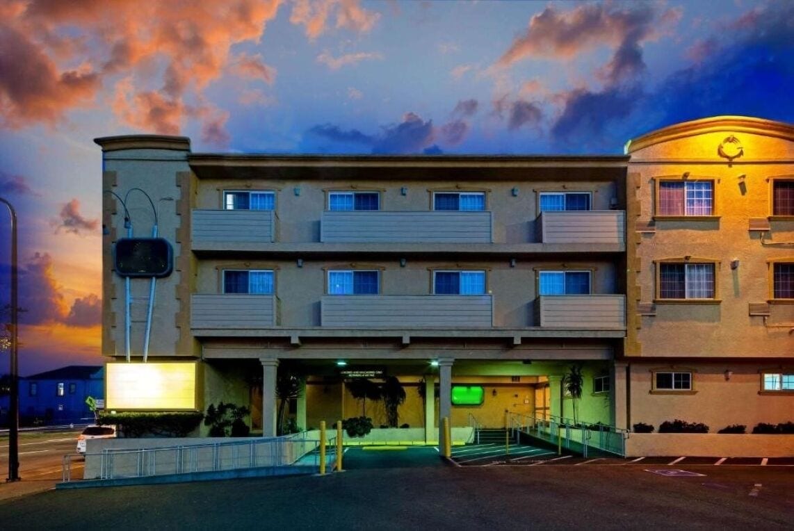 A multi-story hotel with a flat facade, balconies on each floor, and a green-lit entrance.