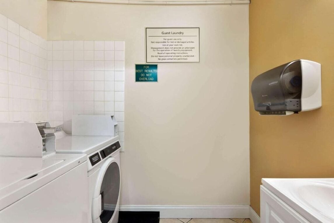 A hotel laundry room with a washing machine, dryer, and a wall-mounted hand dryer. There is a sign on the wall titled "Guest Laundry" with instructions for use.
