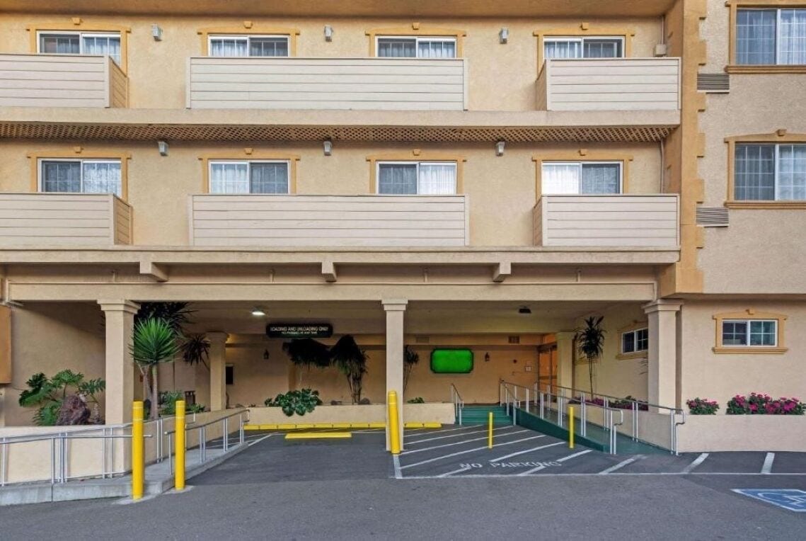 Un hotel de varios pisos con fachada color beige, balcones en cada planta y un estacionamiento delimitado con bolardos amarillos.
