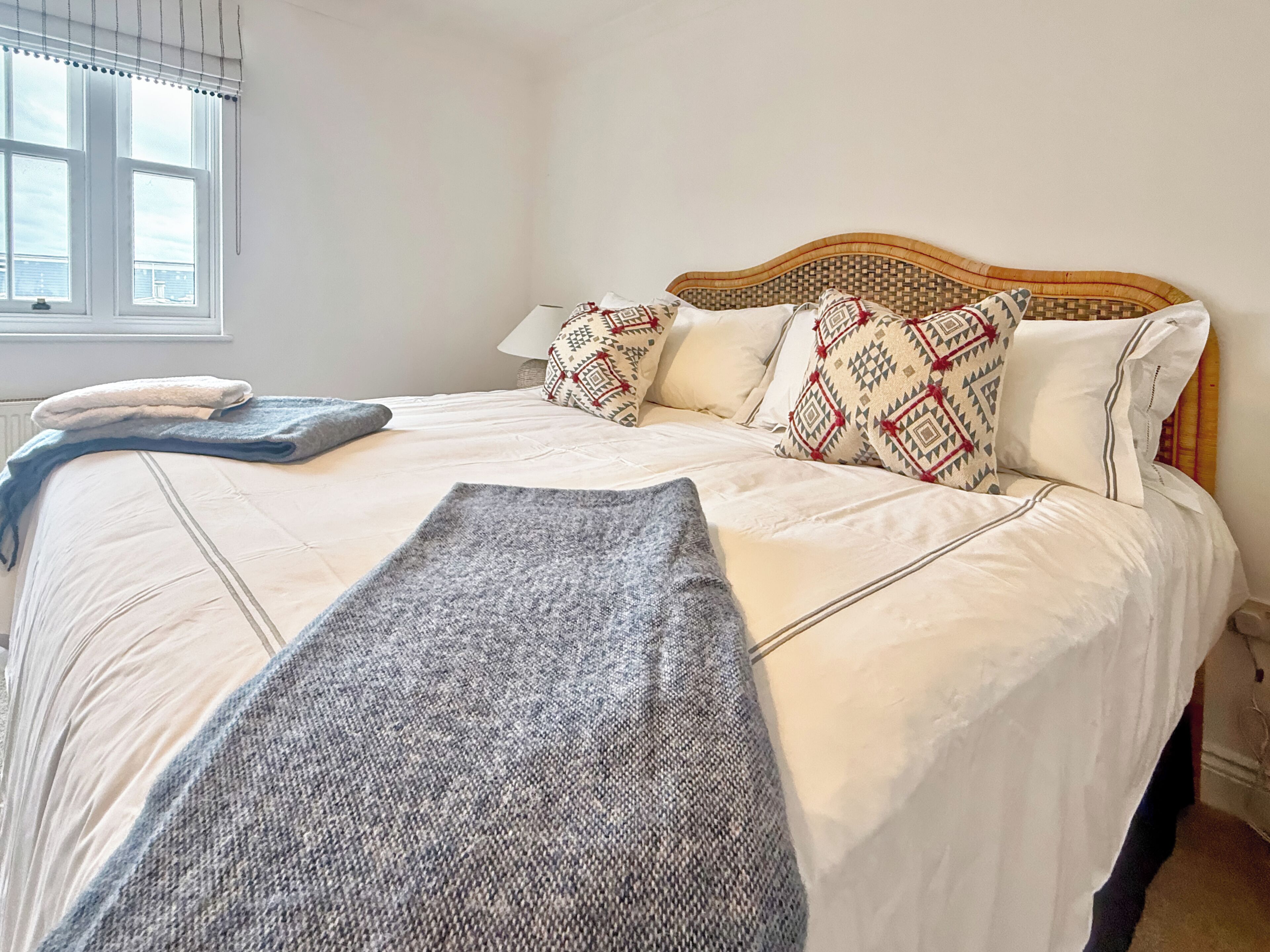A neatly made bed with a patterned headboard, a window with blinds, and a bedside table with a lamp.