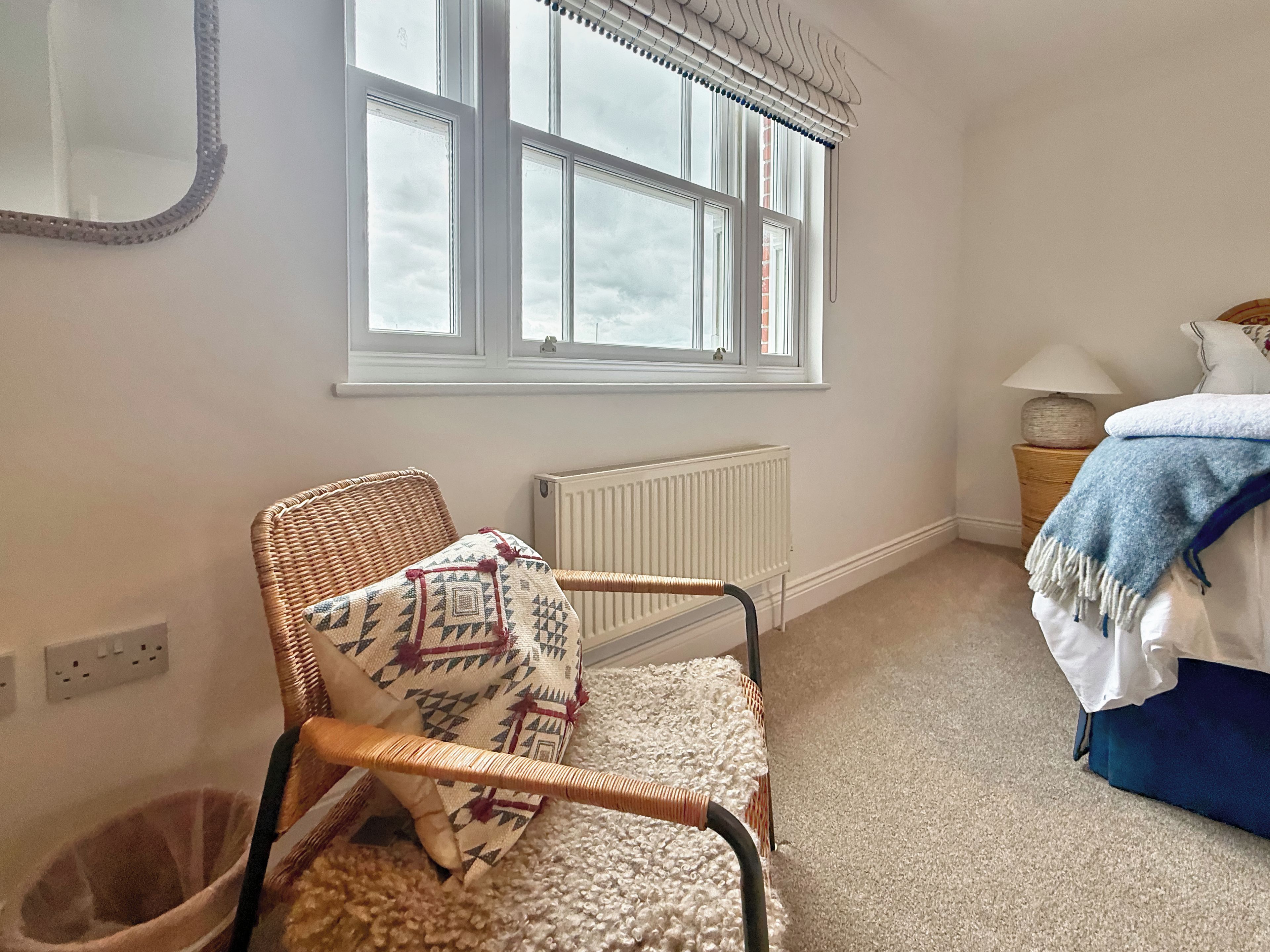 A room with a wicker chair, a radiator, a window with blinds, a bedside table with a lamp, and a bed with a blue blanket.