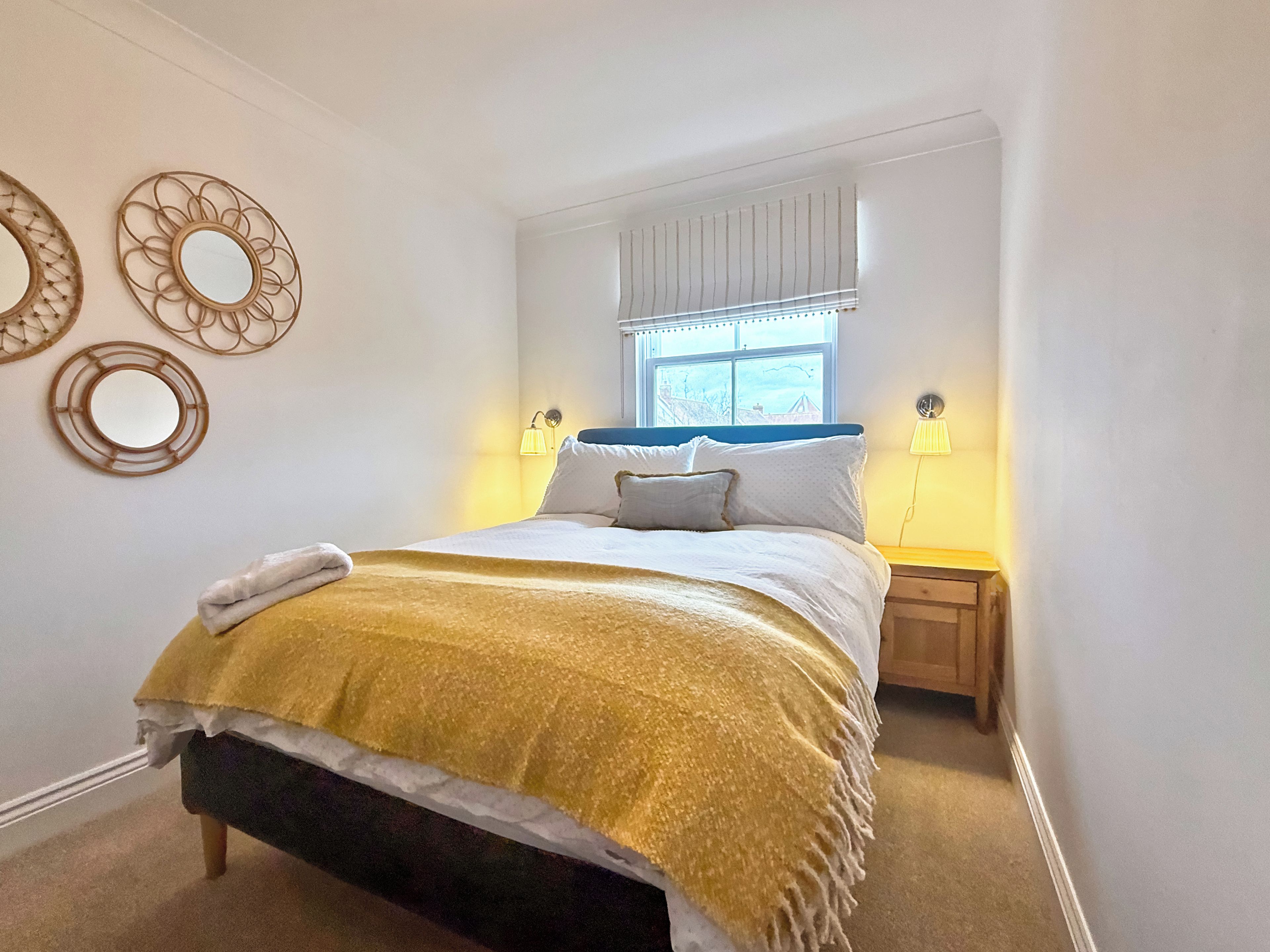 A neatly made bed with a yellow throw blanket, two bedside lamps, and a window with blinds.