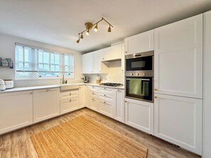 Une cuisine moderne avec des meubles blancs, un four encastré, une cuisinière à gaz et une fenêtre équipée de stores.