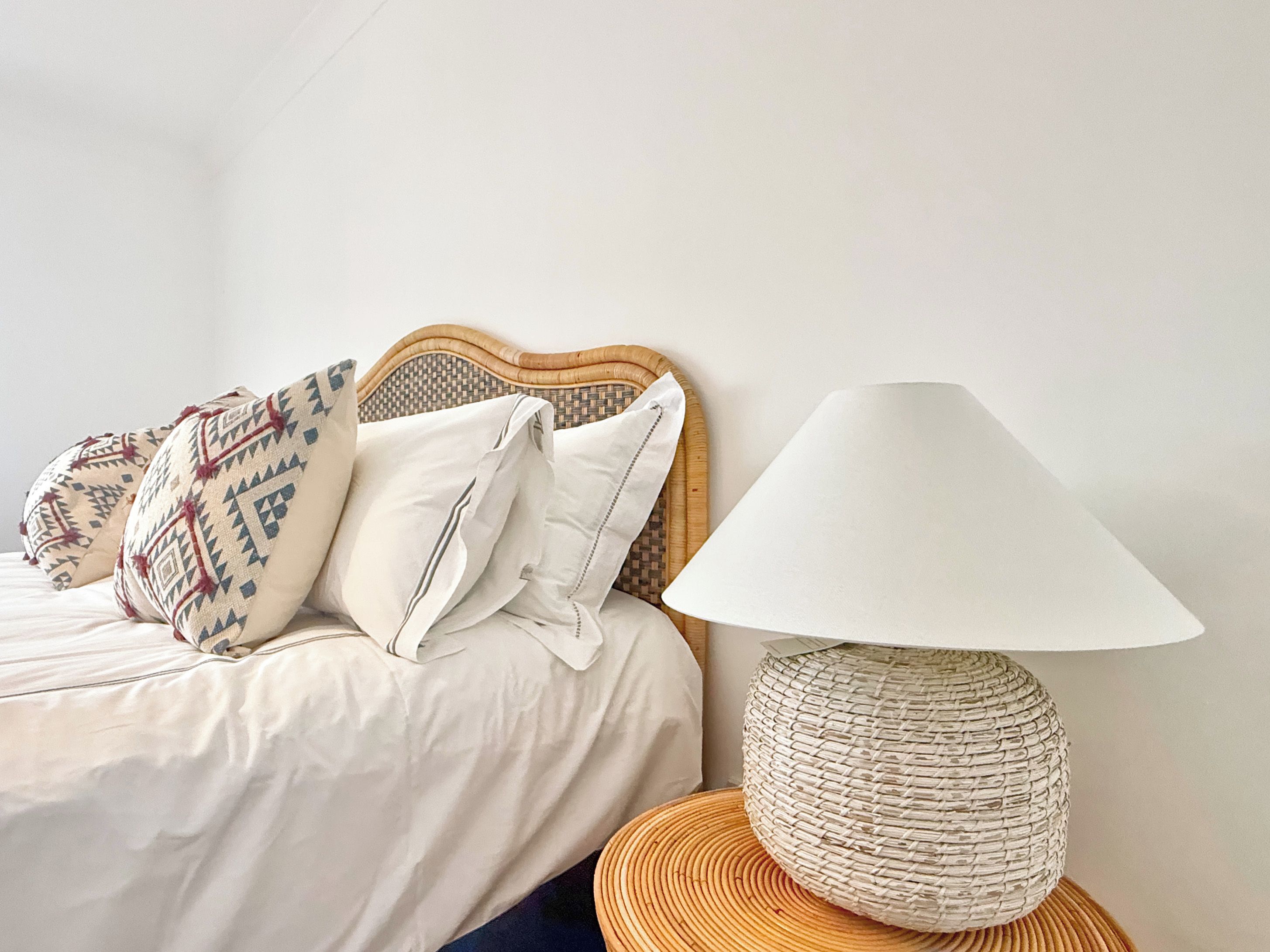 A bed with a woven headboard, a white pillow, and a woven lamp on a round wooden table.