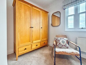 Une pièce comprenant une grande armoire en bois, une chaise en osier avec des coussins à motifs, un miroir au mur et une fenêtre avec des stores.