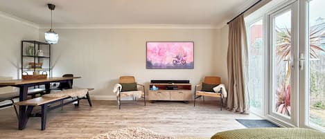 A spacious room with a wooden floor, a dining table, two chairs, a sofa, a television, and a large window with curtains.