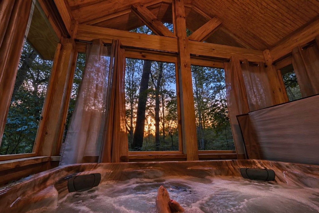 A wooden cabin with a hot tub and large windows overlooking a forest.