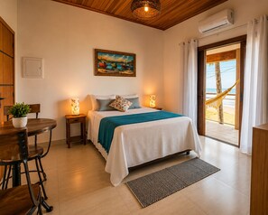 Un dormitorio con cama, lámparas de noche, un cuadro, vista a la playa y una zona de comedor con mesa y sillas.