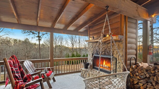 A wooden deck with a stone fireplace, a hanging chair, and a stack of firewood.