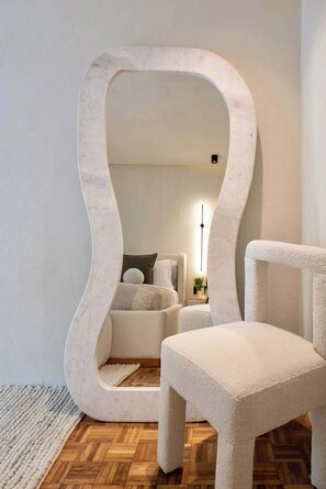 Ein Zimmer mit einem großen, abstrakten Spiegel, einem weißen Stuhl, einem Bett mit Kissen und Holzboden.