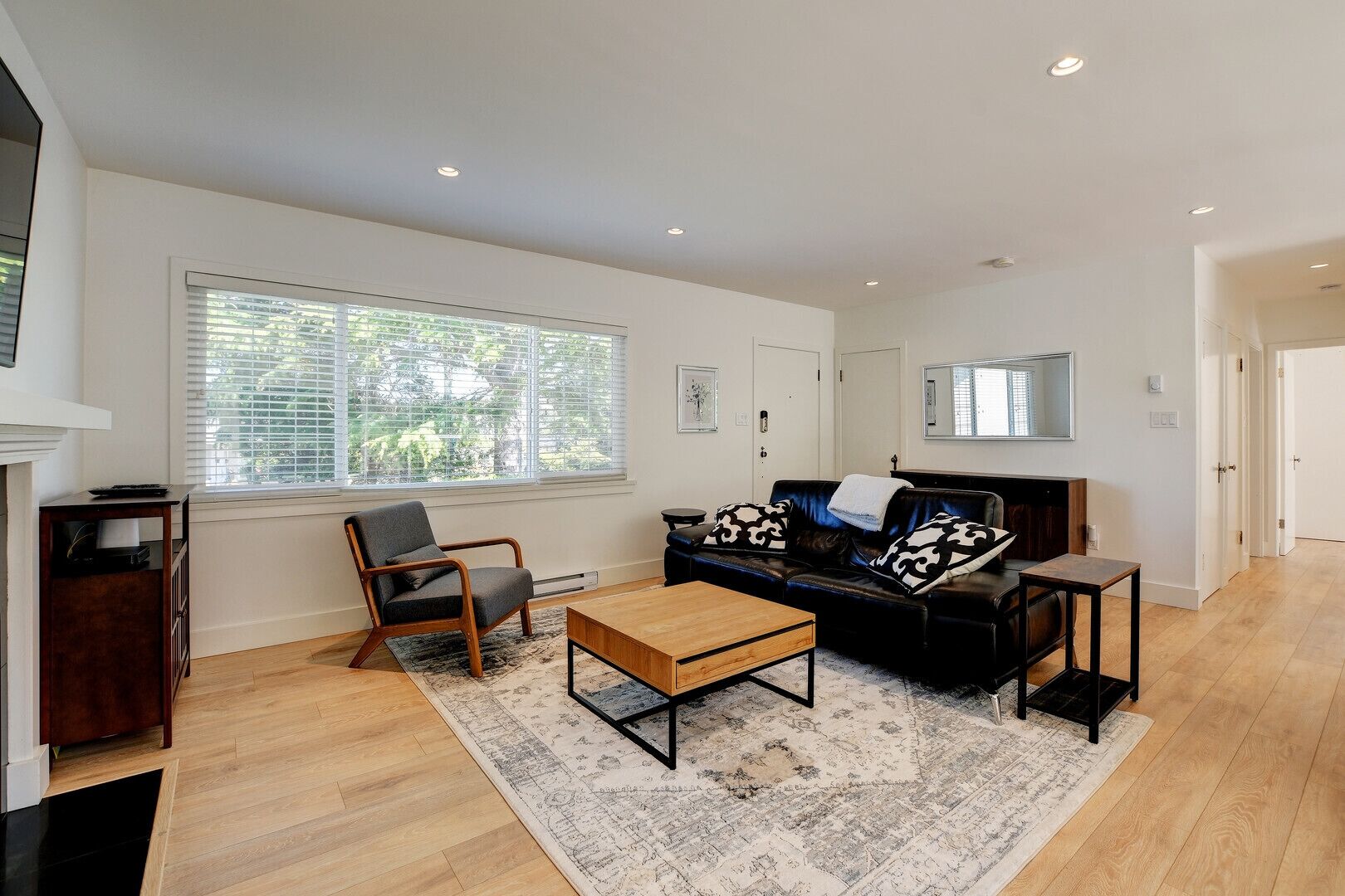 Un salon moderne avec un canapé en cuir noir, une table basse en bois et un fauteuil gris. Il y a une grande fenêtre avec des stores, une cheminée et un miroir au mur.