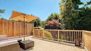 Een terras buiten met een houten terras, een zithoek met kussens, een grote parasol en een houten omheining.
