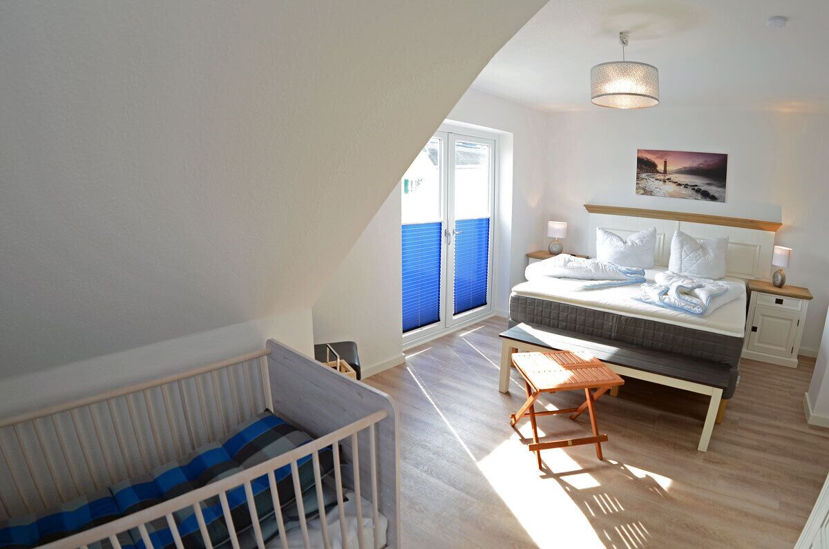 Une chambre à coucher comprenant un lit, des tables de chevet, un banc, un berceau et une fenêtre avec des stores.