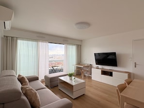 A modern living room with a sofa, coffee table, TV, and a view of the city through large windows.
