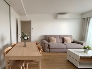 A modern living room with a wooden dining table, beige sofa, and a white side table with a plant.