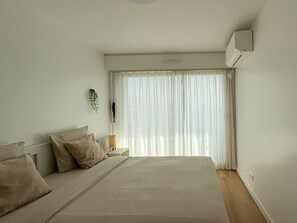 Un dormitorio con cama, mesita de noche, lámpara y ventana con cortinas traslúcidas.