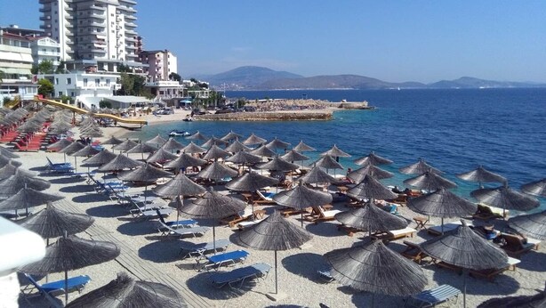 A beach with numerous thatched umbrellas and lounge chairs, a pier extending into the sea, and a backdrop of buildings and mountains. - Alexis Beach (Pet Friendly) (Sarandë)