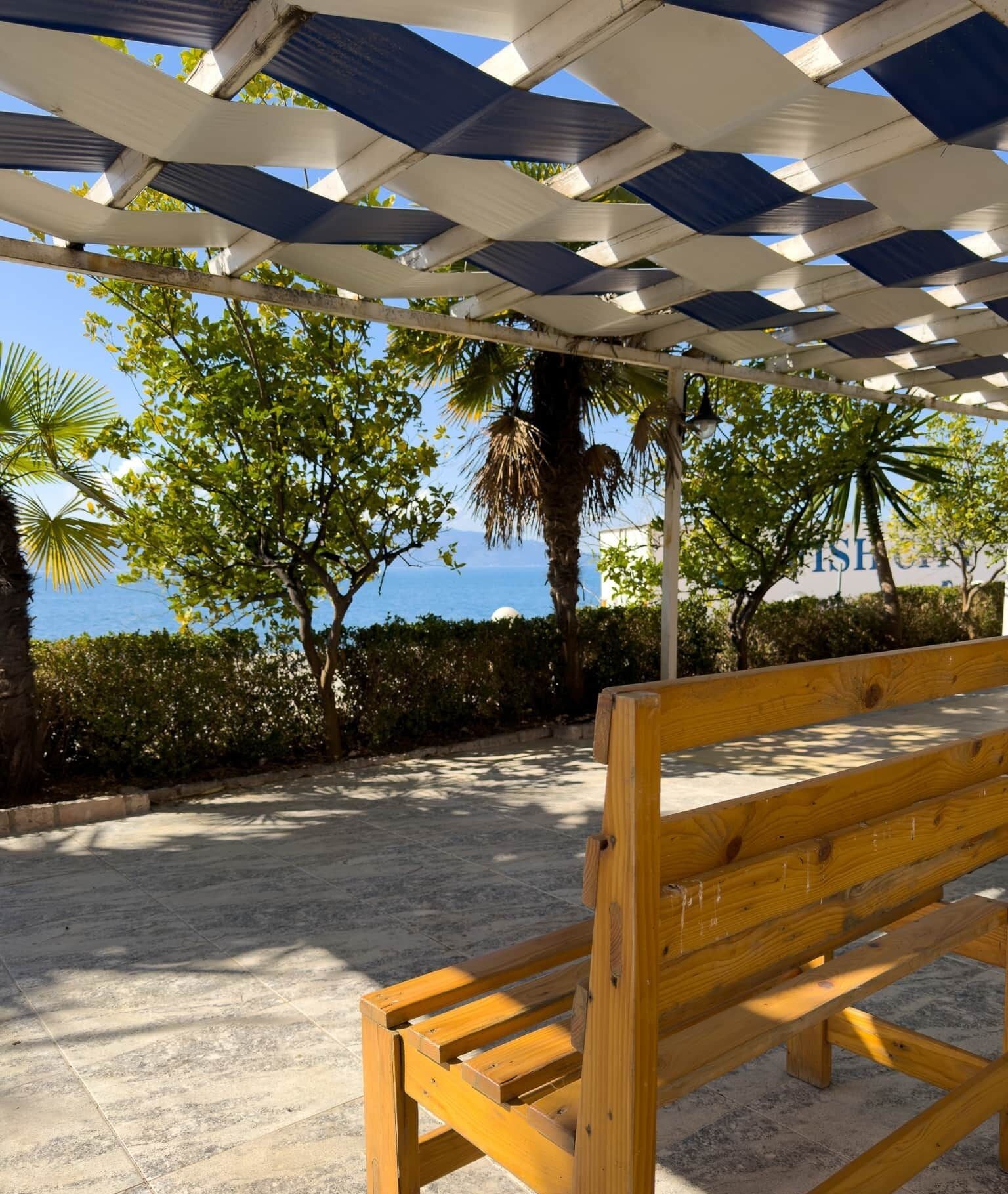 Un banc en bois, placé sous une pergola ombragée, d’où l’on peut admirer la mer et les palmiers.