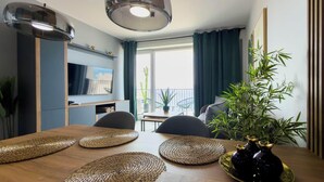 Un coin repas avec une table en bois, des sets de table en fibres tressées, un vase noir et un téléviseur fixé au mur.