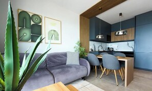 A modern living room with a sofa, dining table, chairs, and a kitchen area with cabinets and a hanging light fixture. - Covista - Panoramic View (Saranda)