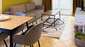 Een moderne woonkamer met een bank, een eettafel en stoelen. Er is een groot raam met gordijnen, een hanglamp en een kleine plant op de vensterbank.